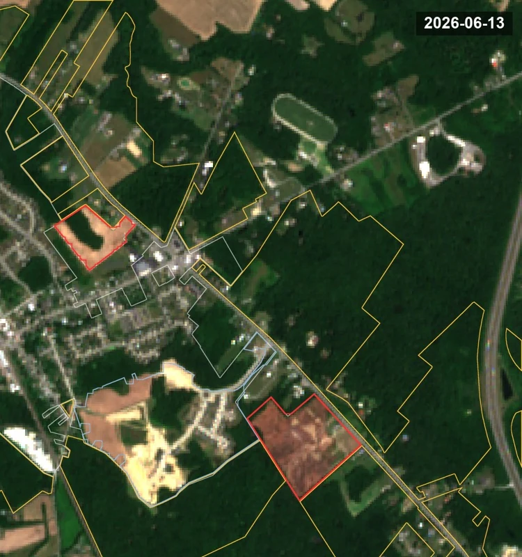 Aerial image dated 13 June 2026. Both red-outlined parcels are bare graded soil, a large subdivision has been cleared south-west of the town center, and dense unbroken woodland still fills the eastern half of the frame.